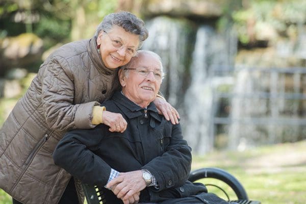 An elderly woman stands behind an elderly man in a wheelchair, both smiling and enjoying a peaceful outdoor setting with greenery and a waterfall—made accessible by 101 Mobility Ramps designed for residential use.