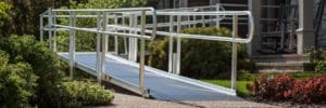 A metal wheelchair ramp with handrails leads up to the entrance of a house, surrounded by green bushes and plants on a sunny day.