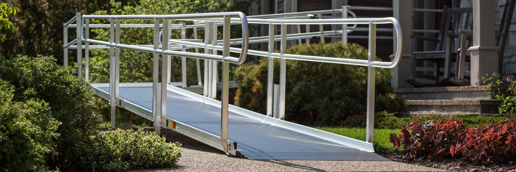 A metal wheelchair ramp with handrails leads up to the entrance of a house, surrounded by green bushes and plants on a sunny day.