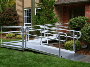 A silver metal wheelchair ramp with railings leads up to the entrance of a house, surrounded by green grass, shrubs, and trees in a landscaped yard.