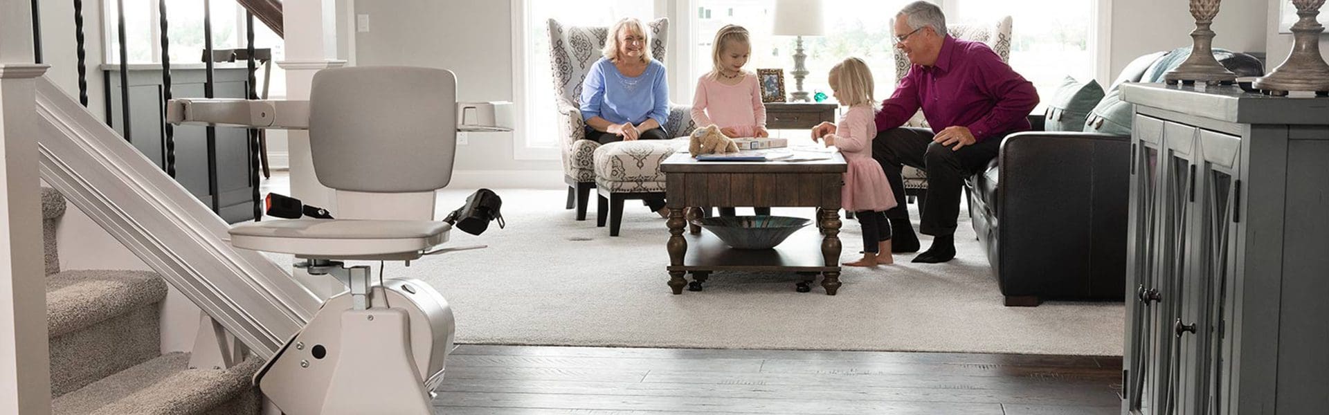 A stairlift is installed next to a staircase in a living room, while two older adults and two young children sit and play together at a coffee table in the background.
