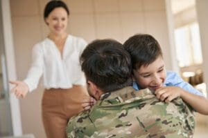 A young boy hugs a person in military uniform, smiling with joy, while a woman in the background extends her arms in a welcoming gesture.