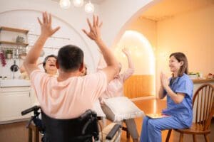 A group of older adults and a caregiver in scrubs are sitting in a bright room, raising their arms and smiling together. One person is in a wheelchair, and everyone appears engaged in a joyful group activity.