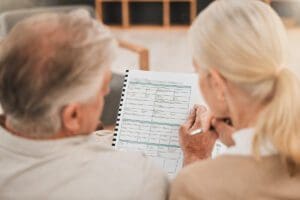An elderly couple sits closely together, reviewing a document with multiple sections and charts, possibly related to finances or healthcare, viewed from behind.