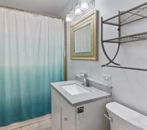 A small bathroom with a white vanity and sink, a mirror with a gold frame, a toilet, metal wall shelves, and a shower with a blue and white ombre curtain under three-light fixture.
