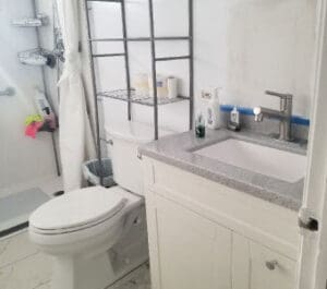 A clean, modern bathroom with a white toilet, metal shelving, white sink vanity, gray countertop, and various toiletries. A shower area with a curtain and cleaning supplies is visible in the background.