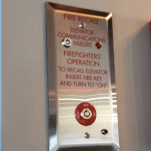 A metal fire recall panel for an elevator with red text instructions, a keyhole, and indicator lights, mounted on a wall for use by firefighters during emergencies.