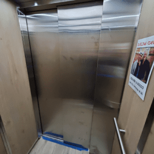 A stainless steel elevator door is partially open inside a wood-paneled elevator. A handrail is visible on the right, and a poster with two people is attached to the wall next to the door.