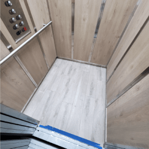 A small, empty elevator with light wood panel walls and a gray wood-patterned floor. The control panel with buttons is on the left, and the elevator doors are partly open.
