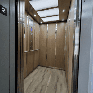 An empty elevator with wood-paneled walls and floor, ceiling lights, and mirrored panels. The elevator is on the second floor, indicated by a number 2 sign near the door.