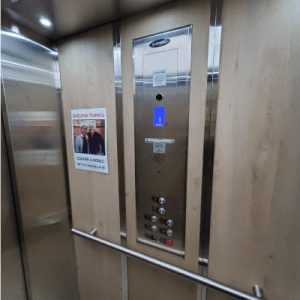 Interior of an elevator with wooden walls, a control panel displaying a blue arrow pointing up, several buttons, a mirror, and a poster on the left wall featuring two people and some text.