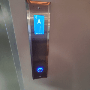 A close-up of an elevator panel with a blue illuminated arrow pointing up and a blue lit button below, indicating the elevator is going up.