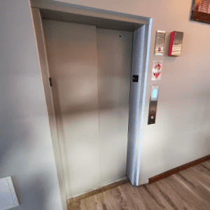 A closed elevator door in a hallway with wall-mounted elevator controls, fire safety signs, and a wooden floor. The elevator call button is illuminated.