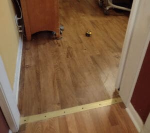 A doorway with a wooden floor and a brass threshold strip. A tape measure is lying on the floor near a wooden cabinet and some exposed electrical outlets are visible on the left wall.