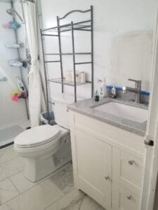A bathroom with a white toilet, metal shelving above it holding toiletries, a white vanity with a gray countertop, sink, soap, and faucet, and a walk-in shower with shelves visible in the background.