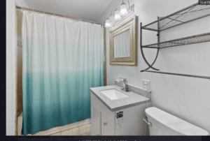A modern bathroom with a blue ombre shower curtain, a white vanity with a gray countertop, a wall-mounted mirror, metal shelves above the toilet, and overhead lights.