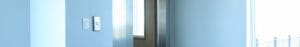 A modern elevator with closed silver doors is set within a light blue hallway. Sunlight streams through a large window on the right, creating a bright and clean atmosphere.