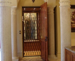A residential elevator with an accordion-style safety gate is visible through an open wooden door, framed by ornate stone columns and decorative arches inside a home.