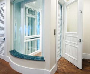 A modern home elevator with glass walls and a white framed door, located in a corner of a room with wooden floors and white walls. A blue-green marble ledge runs along the curved window beside the elevator.