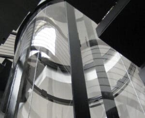 A close-up view of a modern glass elevator shaft with curved transparent walls and black vertical supports, reflecting light from a nearby window above.