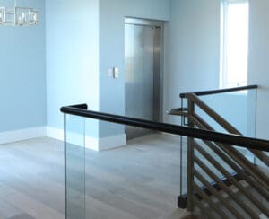 A modern indoor hallway with light wood floors, blue walls, a stainless steel elevator, a window, and a staircase with glass and metal railings.