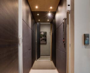 A modern, narrow hallway with dark walls, recessed ceiling lights, and a sliding door. At the end of the hall, there is a framed photo on the wall, and control panels are mounted near the entrance.