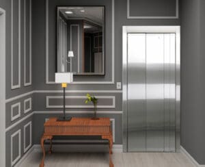 A modern hallway with gray paneled walls, a large mirror, a metallic elevator door, and a wooden table decorated with a lamp and a small vase of yellow flowers.