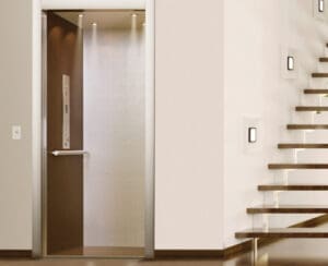 A modern residential elevator with an open door is next to a staircase with wooden steps and glass railings in a well-lit, contemporary interior.