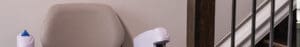 A stair lift chair with armrests and a beige seat is positioned at the bottom of a staircase with metal railings.