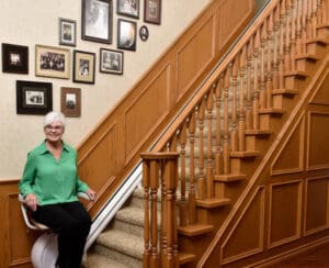 An older woman in a green blouse sits on a stairlift at the base of a carpeted staircase with wood paneling. Framed family photos decorate the wall alongside the stairs.