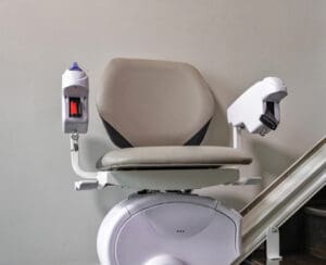 A stairlift chair with padded seat and armrests, positioned at the base of a staircase rail, designed to assist people with mobility challenges in moving up and down stairs.