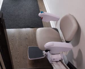 A beige stairlift chair with armrests and a footrest is mounted on a rail along a wooden staircase, next to a white wall and a black floor mat.