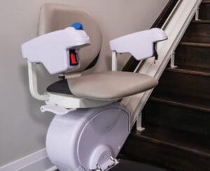 A stair lift chair with padded seat and armrests is installed along a staircase, designed to assist people with mobility challenges in moving up and down the stairs.