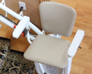 A beige stair lift chair with white armrests is positioned next to a staircase with a patterned carpet and wooden floor.