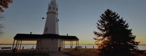 A white lighthouse with a small shelter stands by a large evergreen tree near a lake at sunset, casting long shadows—reflecting the dedication to Neenah accessibility and comfort found in all Home Elevators Neenah solutions.