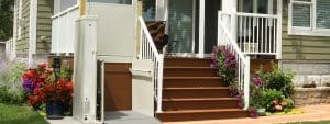 A residential outdoor staircase with white railings, brown steps, colorful potted flowers, and a vertical platform lift for wheelchair accessibility next to the stairs.