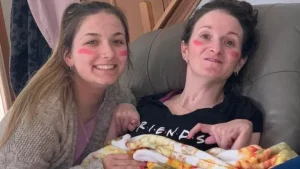 Two women with pink face paint smile while sitting together indoors. One holds a pizza-patterned blanket and wears a black FRIENDS shirt, as they share a moment of joy—showcasing the warmth of the Access Elevator Community spirit.