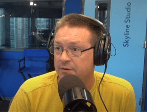 A man wearing glasses and headphones speaks into a microphone in a studio. He is wearing a yellow shirt, and behind him is a blue wall labeled Skyline Studio with windows showing city buildings.