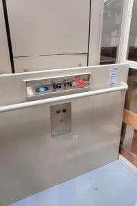 A close-up view of an elevator control panel with buttons, switches, and emergency stop, mounted on a light-colored wall inside an elevator. There are handrails and a wooden barrier visible on the right side.
