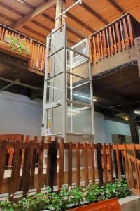 A glass elevator with white framing is located indoors, surrounded by wooden railings and balconies. There are green plants with flowers at the base and wooden beams overhead. The area has a warm, rustic atmosphere.