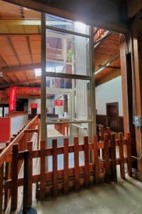 A modern wheelchair lift with glass panels is installed inside a rustic building with wooden railings and beams. There are red signs in the background and natural light enters from above.