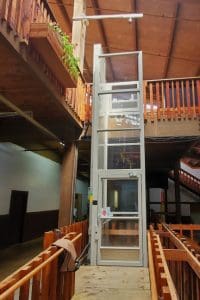 A glass elevator with a metal frame is situated indoors between wooden railings and balconies, providing access to upper floors in a rustic, wood-accented building.