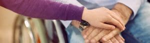 A younger person's hand gently rests on top of an older person's hands, who is sitting in a wheelchair, conveying care and support as part of our Accessibility Projects Community.