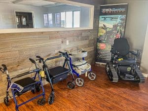 A room with wood walls and floors displays three types of walkers and a black tracked power wheelchair. A TrackMaster Mobility banner stands in the corner next to a large window.