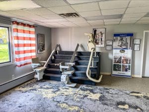 A showroom displays two stair lifts installed on a short set of carpeted stairs, with striped curtains on a window, promotional posters, and a patterned rug on the floor.