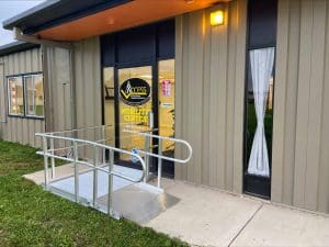 A beige metal building with a glass door labeled Access Mobility Center and a metal wheelchair ramp leading to the entrance. A light is on above the door, and a white curtain covers one window.