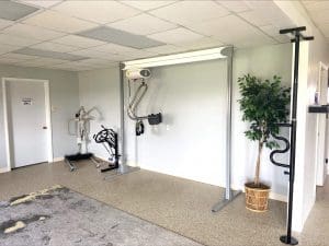 A room with exercise and rehabilitation equipment, including a patient lift, walker, and stationary bike, with a potted plant in the corner and a door labeled Employees Only on the left.