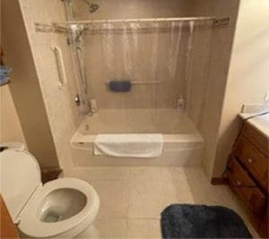 A bathroom with a white toilet, a bathtub with a clear shower curtain, grab bars, a blue bath mat on the floor, a towel draped over the tub, and a wooden vanity with a sink.