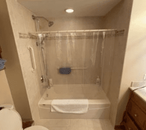 A bathroom with a bathtub-shower combo, a clear shower curtain, a grab bar, a towel on the tub edge, and toiletries on the ledge. Part of a toilet and sink are visible on the left and right sides.