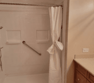 A bathroom with a walk-in shower, a white shower curtain tied in the middle, a grab bar on the shower wall, and a beige countertop with wooden cabinets on the right.